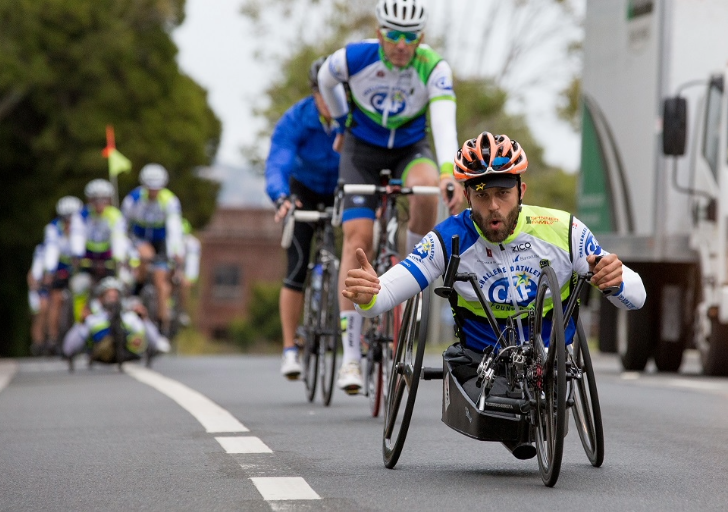 Andre Kajlich leading a group ride in a handcycle, giving a shaka while wearing a helmet and CAF cycling jersey