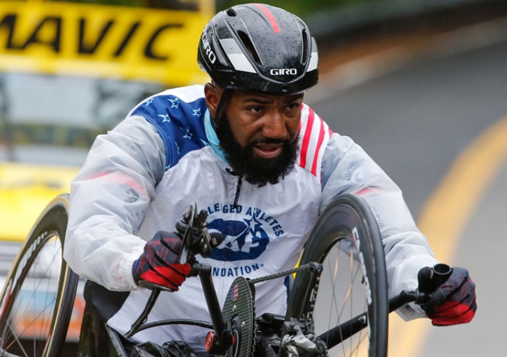 A handcycle athlete wearing a helmet and a CAF racing jacket powers up a road in the rain, gripping the handcycle handles with determination. A bright yellow support vehicle follows behind.