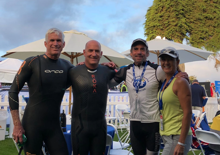 Group of four triathlon participants smiling together after the event, wearing wetsuits and race gear, with umbrellas and chairs set up in the background.