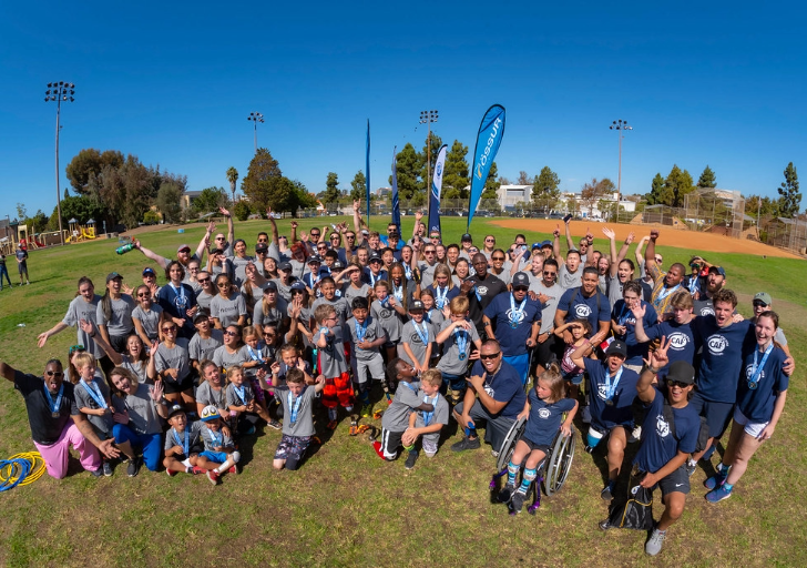 A large group of athletes, families, and volunteers gather on a sunny field during an Össur and CAF mobility clinic, smiling and cheering together.