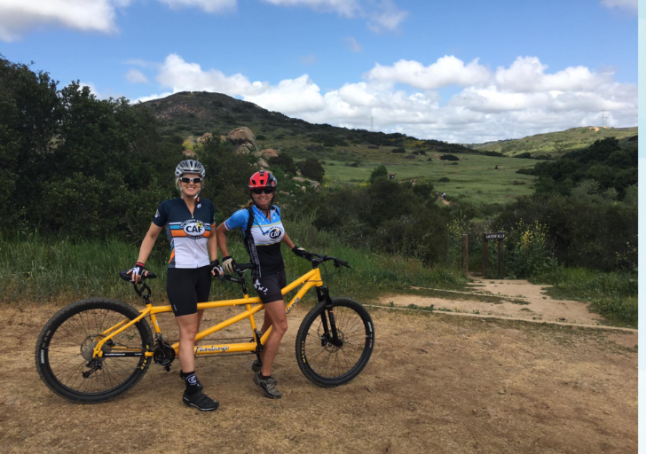 Two athletes with physical disabilities stand smiling beside a yellow tandem bike on a dirt trail, wearing helmets and CAF cycling kits, with rolling green hills and open sky in the background.