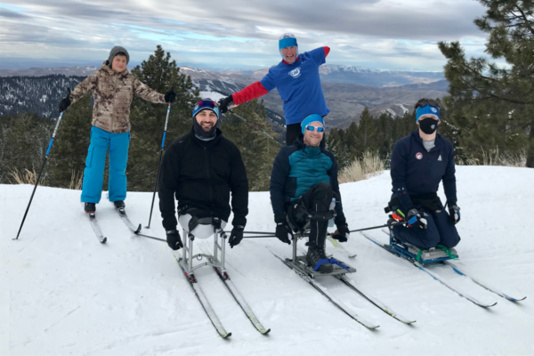 Five athletes with physical disabilities pose on skis at a mountain overlook, smiling and celebrating together with snow-covered peaks and valleys behind them.