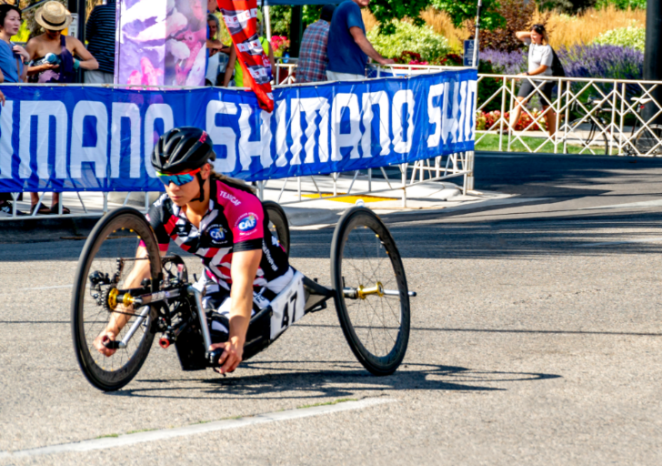 Athletes with physical disabilities compete in a handcycling race, riding low-profile racing handcycles in CAF jerseys as they round a turn on a closed city course.