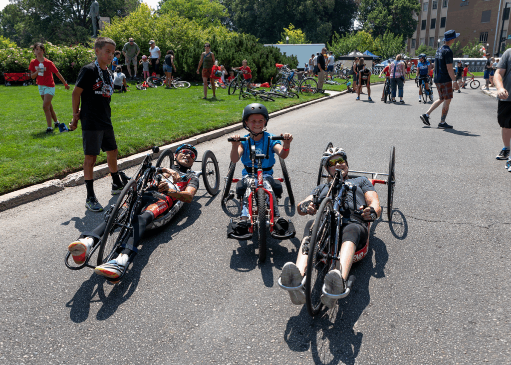 Two adult athletes in handcycles smile alongside a young child riding a smaller adaptive cycle, surrounded by other participants and families at an outdoor cycling event.