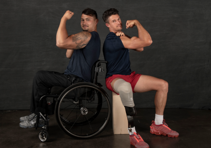 Two athletes pose back-to-back, flexing their arms—one seated in a wheelchair and the other with a prosthetic leg.