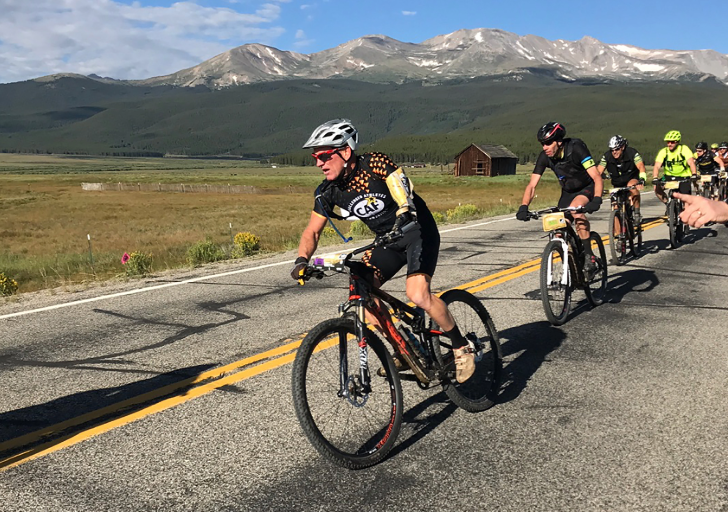 Cyclist wearing a CAF jersey rides on a mountain road during a race, with a scenic backdrop of open fields and mountains.