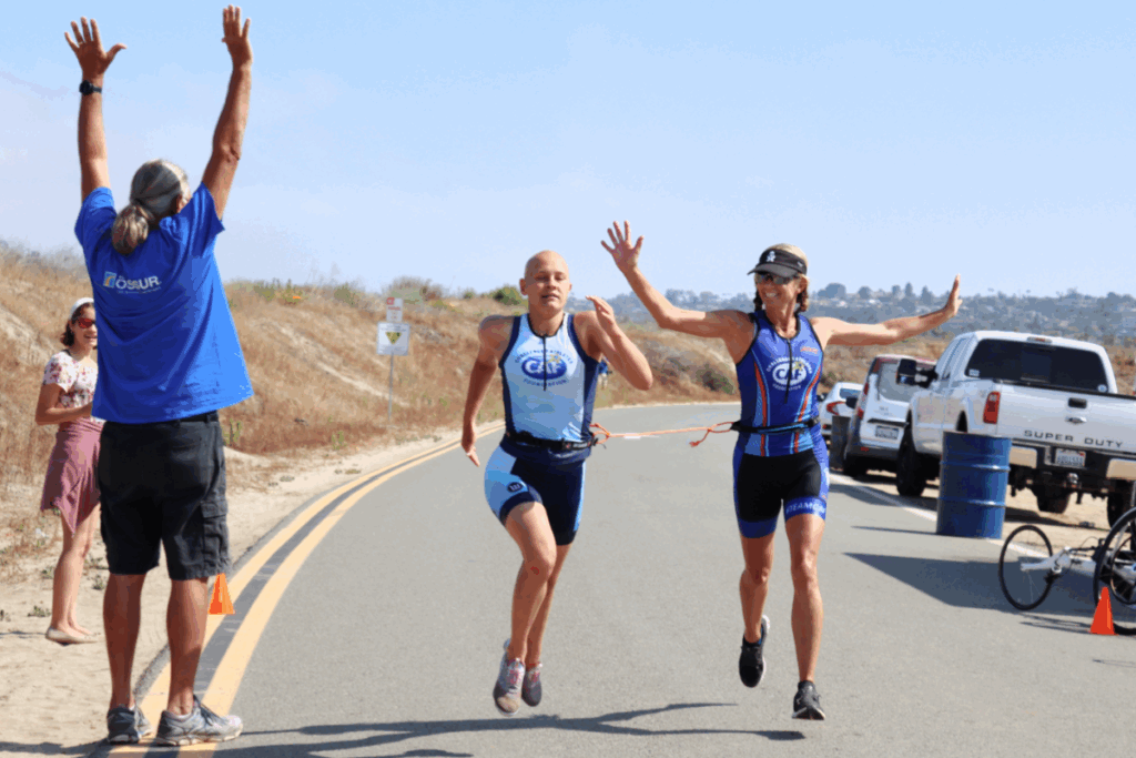 Two triathletes running toward the finish line with their guide, raising their arms in celebration.