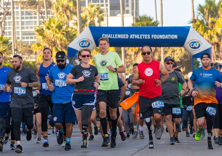 A large group of runners, including athletes with physical disabilities using running prosthetics, take off from the starting line under a Challenged Athletes Foundation archway. Palm trees and tall buildings line the background as the diverse group of participants run together, creating an energetic and united atmosphere.