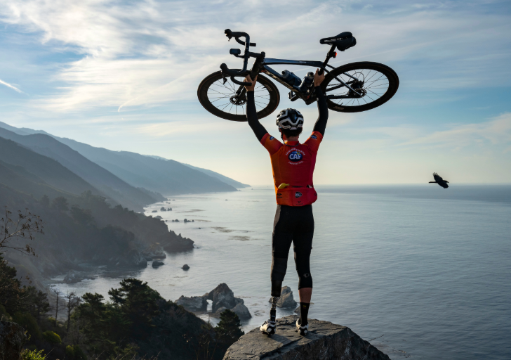 Cyclist wearing a CAF jersey holds their bike overhead on a cliff overlooking the Pacific coastline during Million Dollar Challenge.