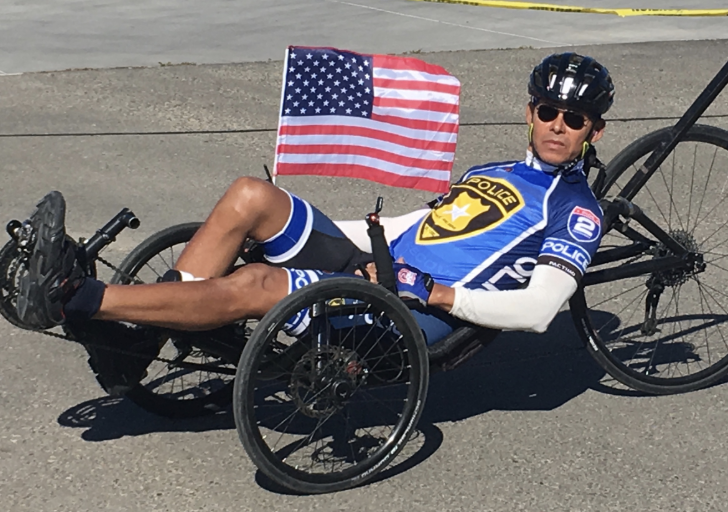 A man with a physical disability rides a recumbent handcycle while wearing a blue police cycling jersey and helmet. An American flag is attached to the back of his cycle, waving in the air. He wears sunglasses and cycles on a paved road in bright sunlight.