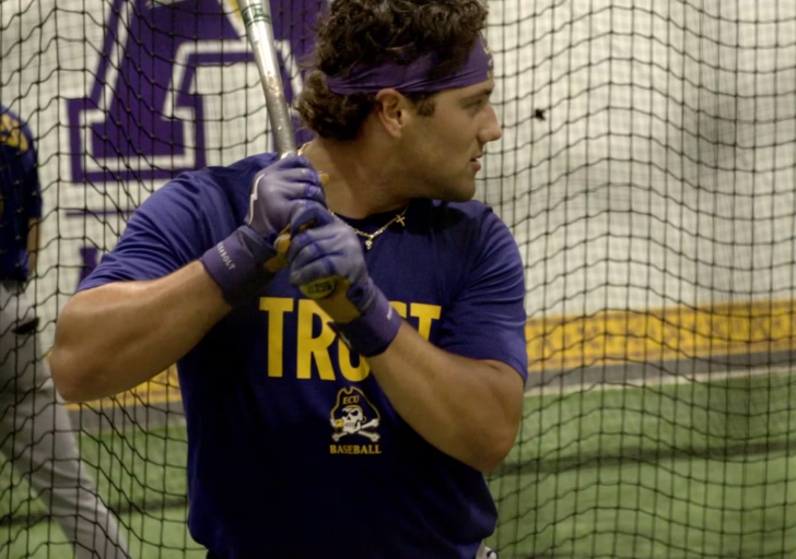 A college baseball player wearing a purple “TRUST” shirt and headband prepares to swing a bat inside a batting cage.