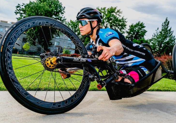 Close-up of an athlete with a physical disability in a racing handcycle, wearing a helmet, sunglasses, and a black-and-blue team jersey. The athlete leans forward with focus, gripping the hand pedals tightly.