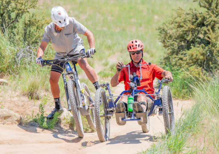 Participant on an adaptive mountain bike navigating rocky terrain with a fellow cyclist riding beside them