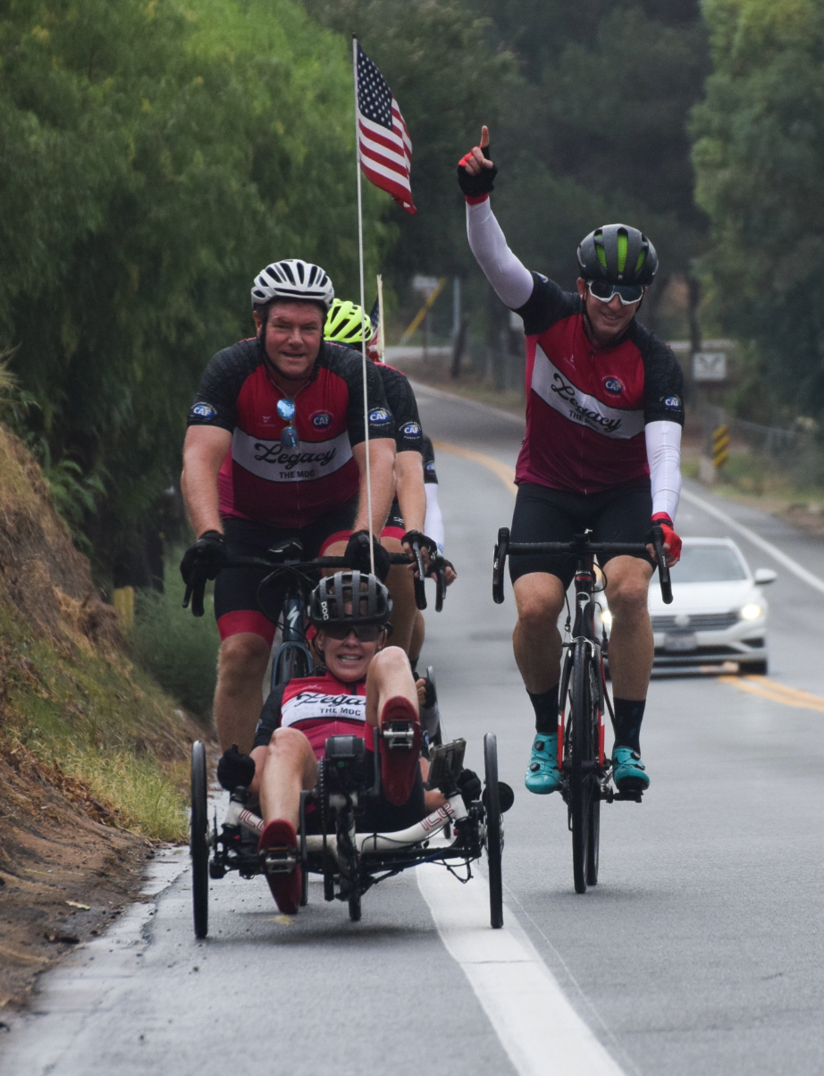 Cyclists and a handcycle athlete ride together on a road, one rider raising a finger as a small American flag waves behind them.