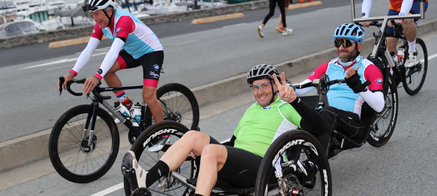 Two athletes ride handcycles alongside a cyclist on a coastal road, smiling and signaling as they move together.