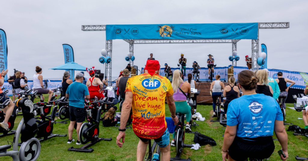 Crowd of cyclists riding stationary bikes on grass facing a stage with event signage reading “Tour de Cove Salty Spin.