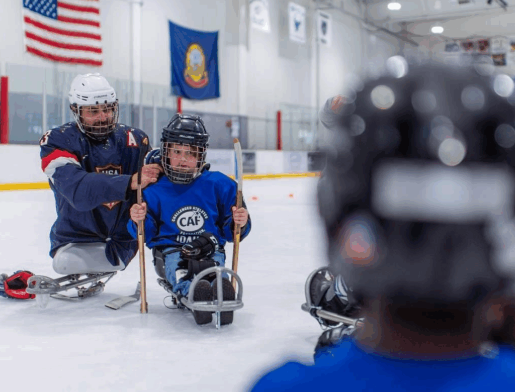 Josh Sweeney in sled hockey sled on ice helping young boy play hockey