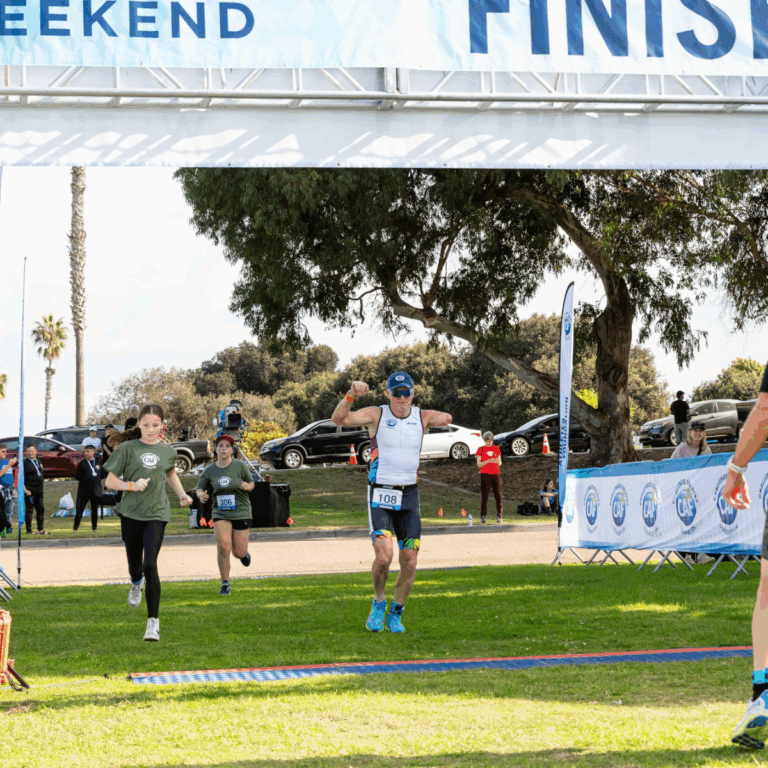 Willie Stewart crosses the finish line of a triathlon with arms raised in celebration, alongside young volunteers in green shirts.