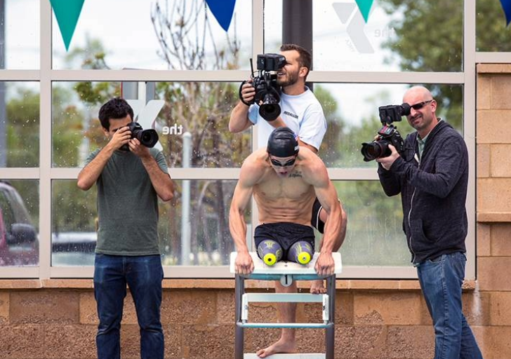 Swimmer with bilateral leg prosthetics prepares to dive from the starting block while three photographers capture the moment poolside.
