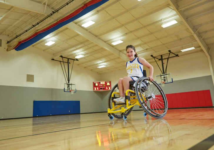 Young athlete in a yellow sports wheelchair smiles on a basketball court wearing a team jersey.
