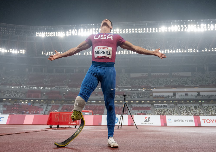 Paralympic sprinter wearing a USA uniform celebrates on the track with arms outstretched at Tokyo Stadium.