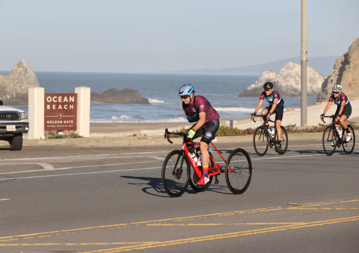 Cyclist rides a red adaptive trike along Ocean Beach with two other cyclists following behind on a sunny day.
