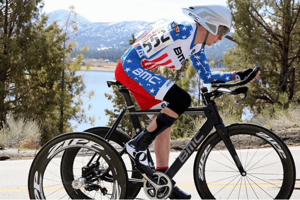 Cyclist wearing a USA skinsuit rides a BMC time trial bike near a lake with snowy mountains in the background.