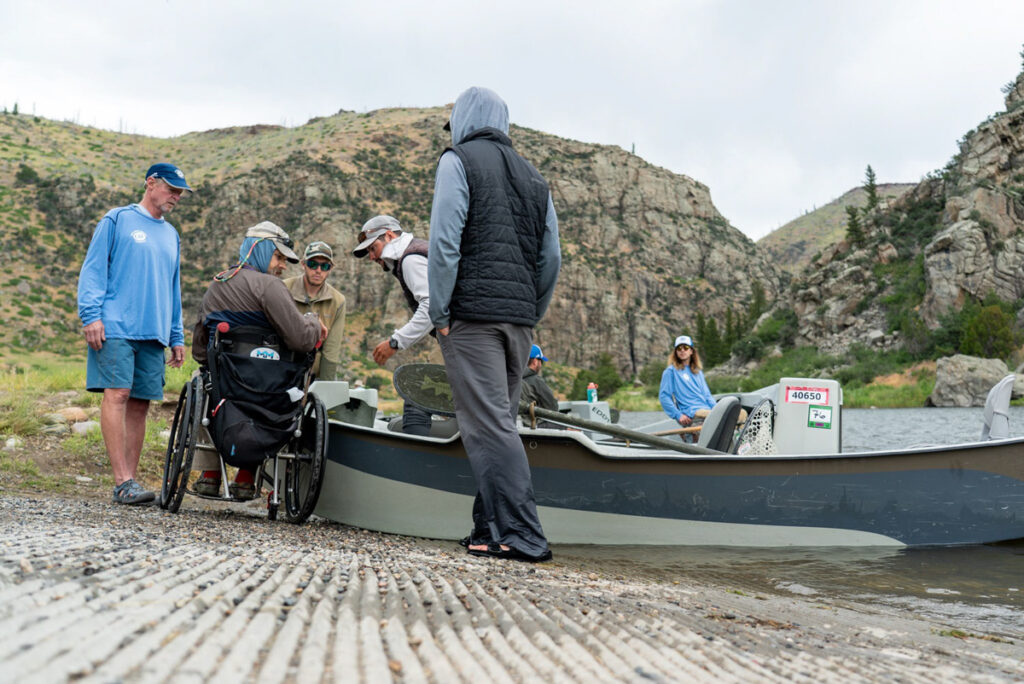 Group assisting a person in a wheelchair onto a fishing boat by a riverbank with mountains in the background.