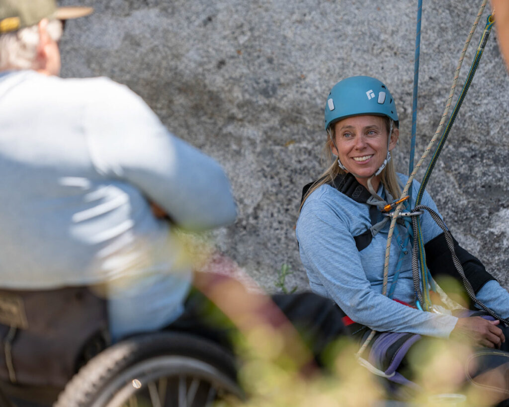 Woman in a helmet smiling at a man in a wheelchair near a rock face, with climbing ropes visible.