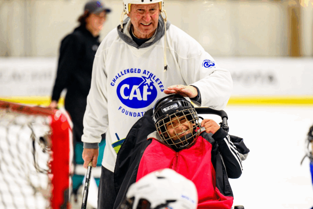 A hockey coach in a white CAF jersey smiles while patting a young sled hockey player wearing a helmet and red bib on the head. The athlete beams with joy on the ice.