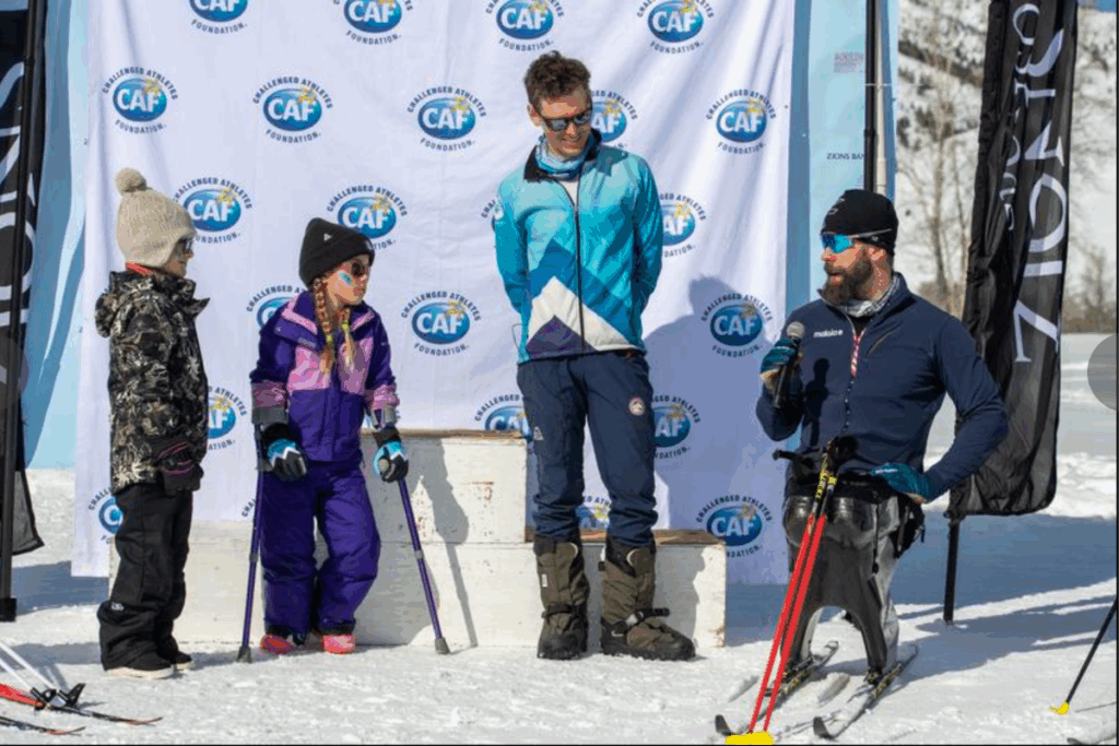 Aaron Pike speaks to a group of young participants at a CAF winter adaptive sports event, with a branded backdrop behind them.