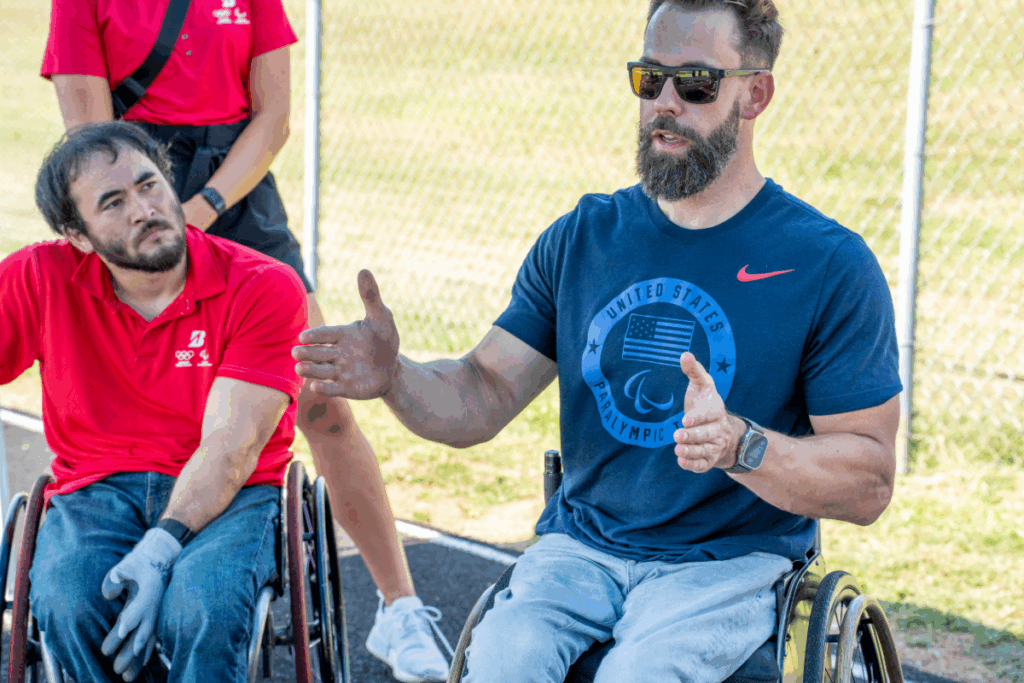 Aaron Pike speaks to a group of athletes during a track event, gesturing with his hands while sitting in a wheelchair next to Chuck Aoki.