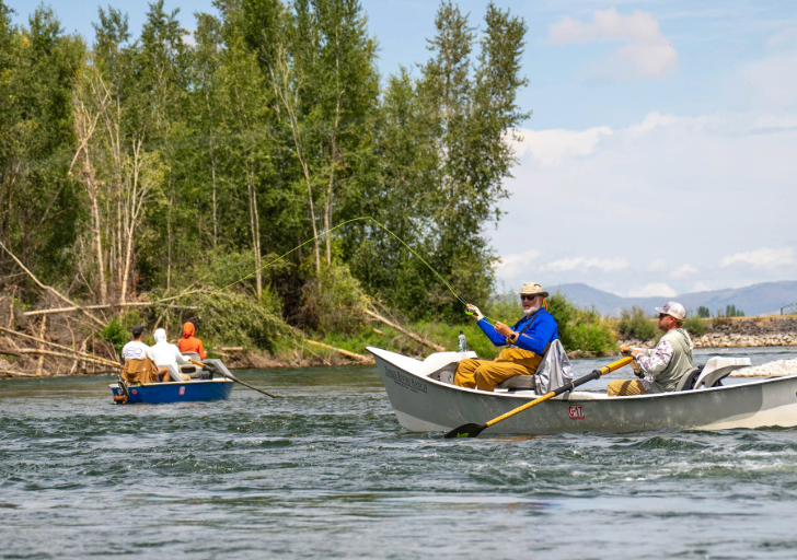 Two people fly fishing in a boat with three people in an adjacent boat on a sunny day.