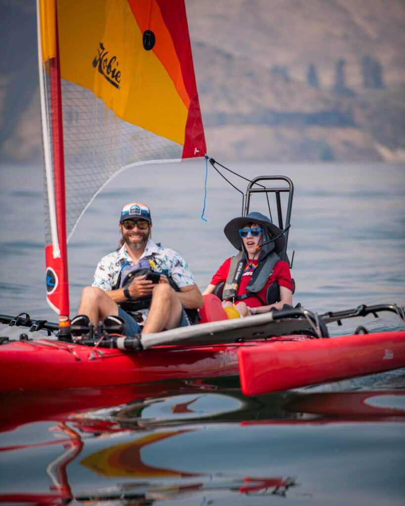 Two people on a red sailing kayak with a colorful sail on calm water.
