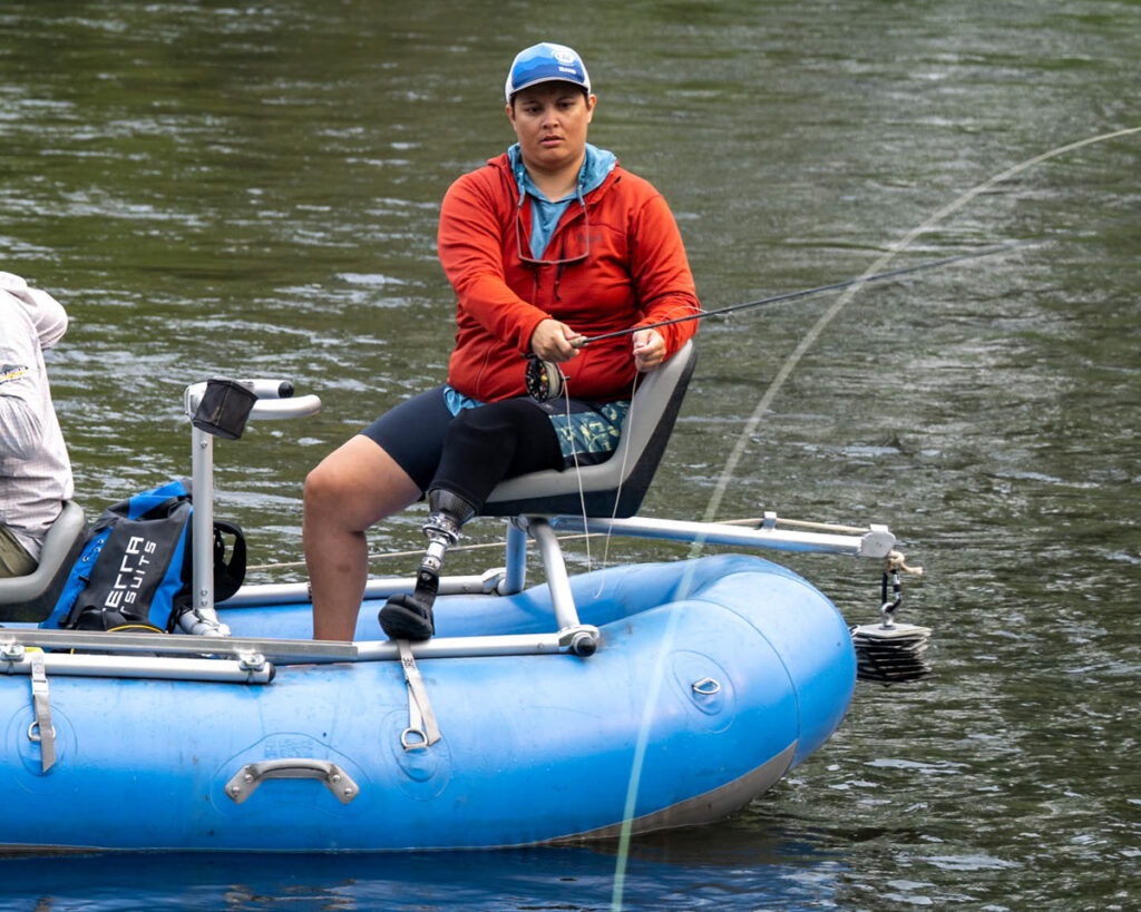 Person fishing from a blue inflatable raft on calm water, wearing a red jacket and prosthetic leg.