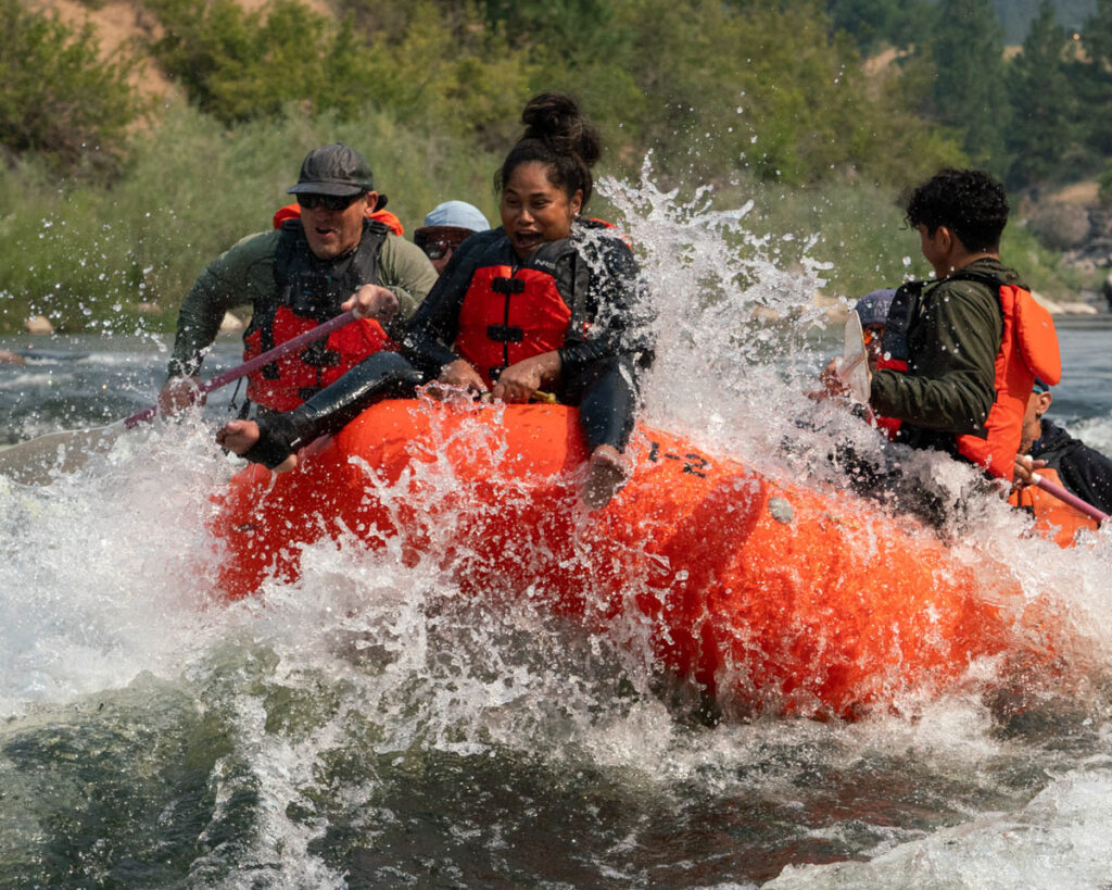 A group of people white-water rafting on a river in an orange raft, surrounded by splashing water.