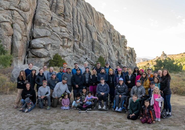 A large group of people posing in front of a tall, ridged rock formation under a partly cloudy sky; several of the people are in wheelchairs.