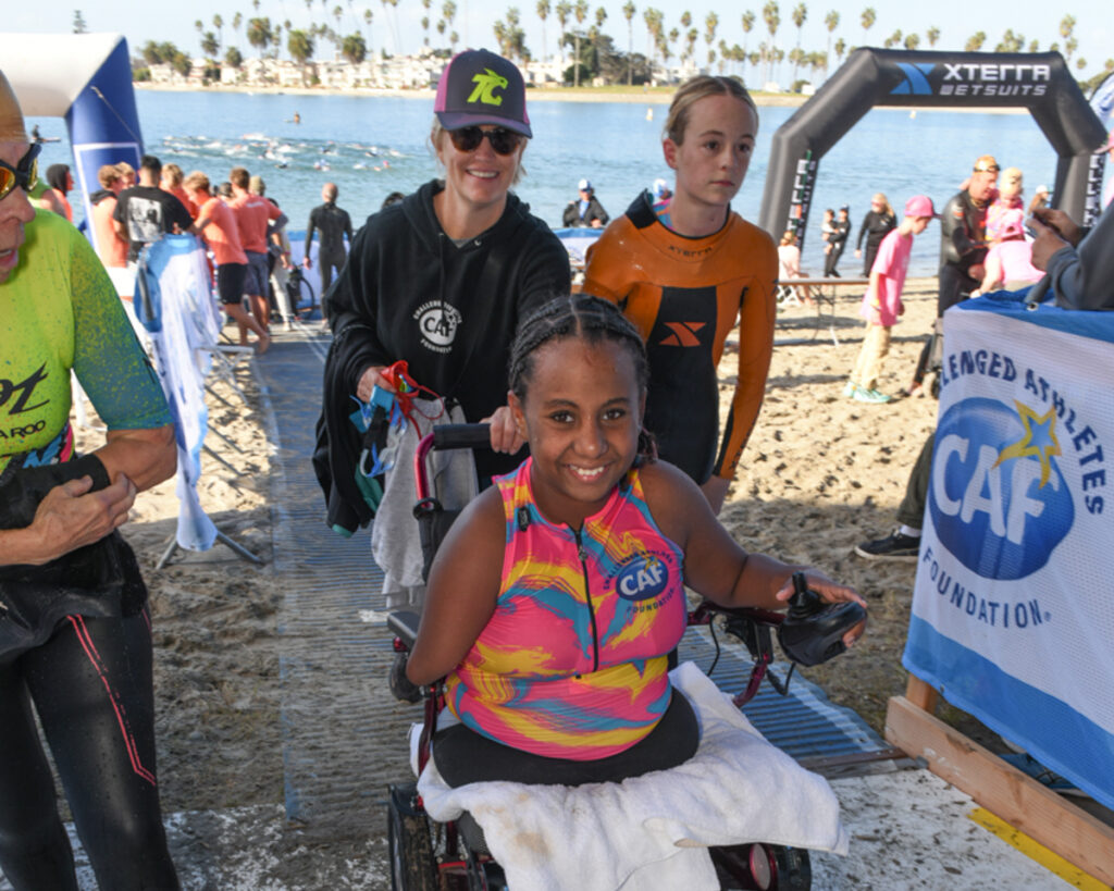 A young person with upper and lower limb differences in a wheelchair smiling at a beach event, with people in wetsuits and a waterfront in the background.