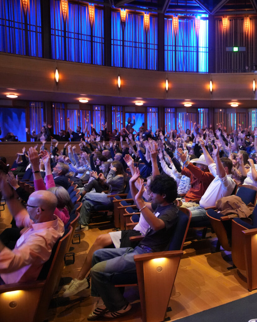 A crowded auditorium with people raising their hands, surrounded by blue and warm yellow lighting.