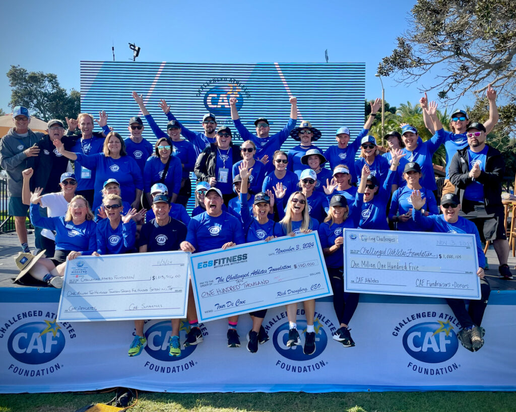 A group of people in matching blue shirts posing with oversized checks in front of a large screen outdoors.