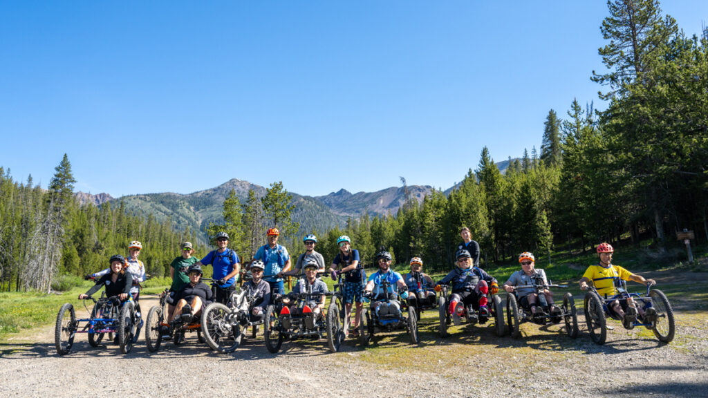 A group of people on adaptive bikes in front of mountains and trees under a clear blue sky.