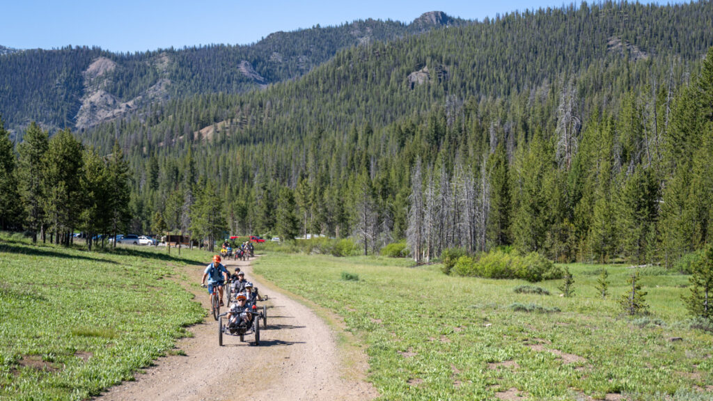 A group of cyclists rides along a dirt path through a grassy field with a forest and mountains in the background.