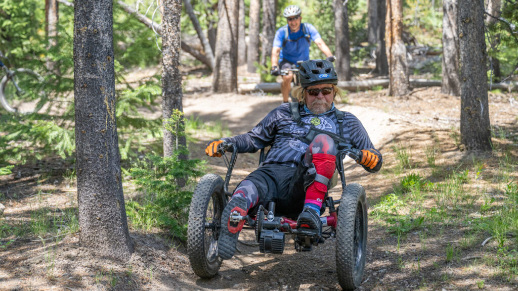 Person riding a recumbent bike on a forest trail, with another cyclist visible in the background.