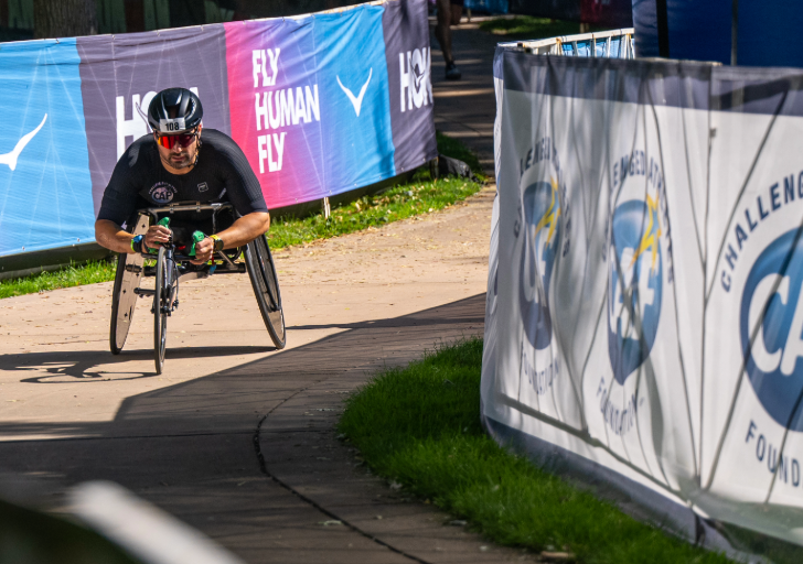 A wheelchair racer rounds a corner on the course, passing banners that read “Fly Human Fly” and “Challenged Athletes Foundation.”
