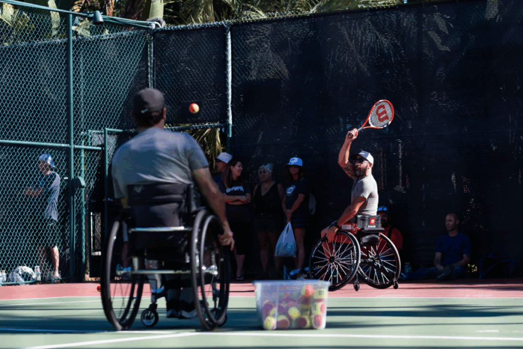 Two athletes in sport wheelchairs rally on the tennis court during an adaptive tennis clinic, with spectators watching.