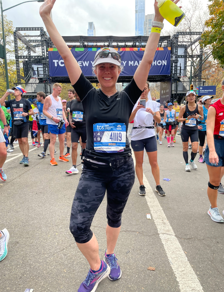 Runner celebrates at the finish line of the TCS New York City Marathon, smiling with arms raised in victory.