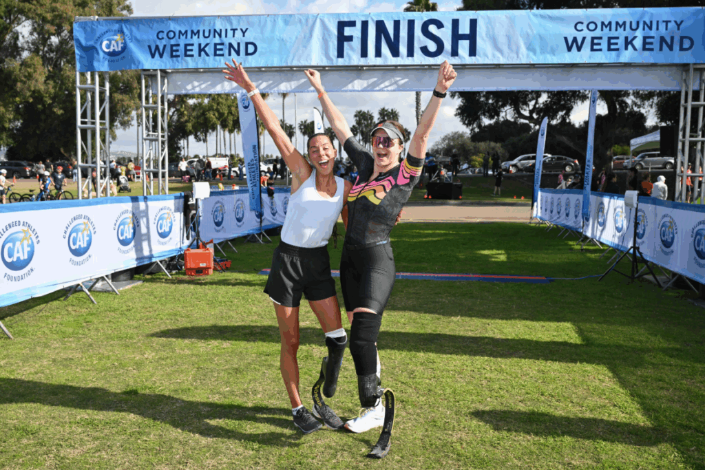 Two athletes with lower limb sports prostheses at the SDTC finish line with hands up in celebratory pose.