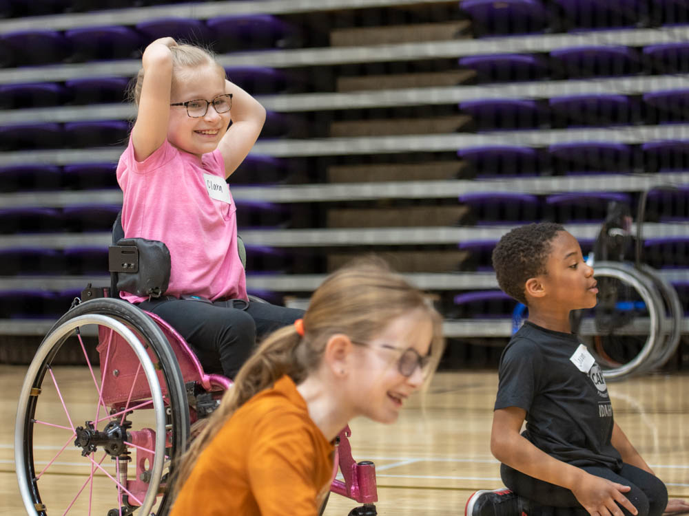 Three children in a gymnasium, one in a pink wheelchair, smiling.