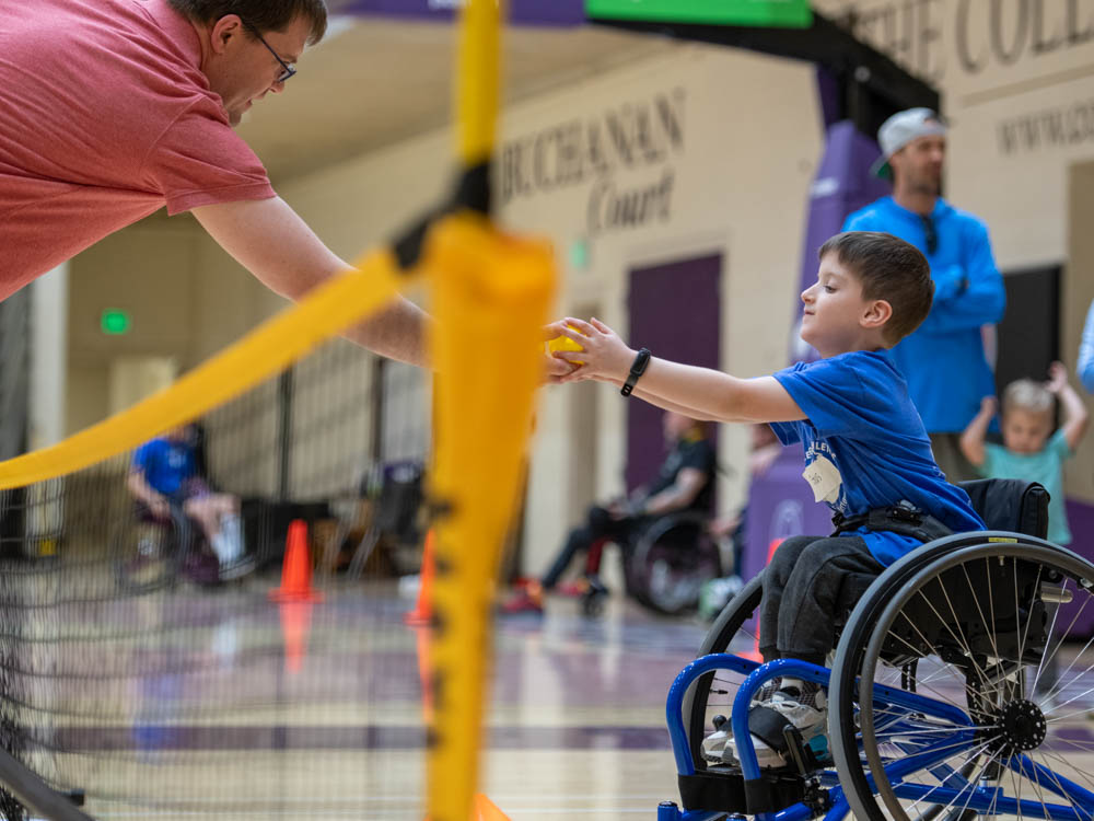 Child in wheelchair exchanging a yellow ball with an adult across a net in a gymnasium.