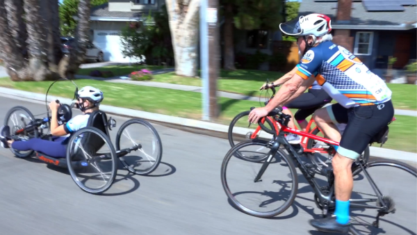 Cyclists, including a handcyclist, ride together on a sunny neighborhood street during Ride for Pie Day.